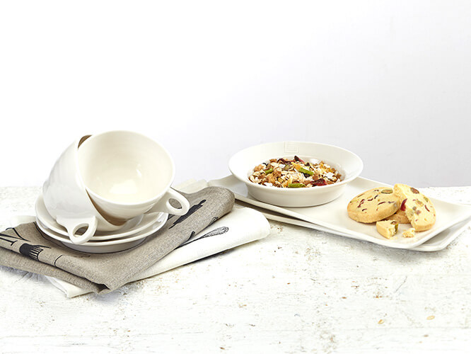 Dining items on a table setting, including ceramic muesli bowls, teacups and linen teatowels.